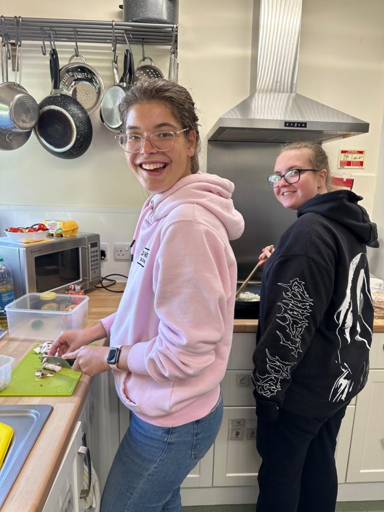 2 People from All Sort'd in a kitchen cooking a meal, one person stood on the left has a big smile on their face while chopping mushrooms and the person behind them is stood at the stove stirring frying onions.